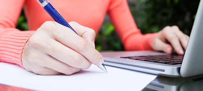 Een vrouw, zittend in een tuin, schrijft met de hand een ontslagbrief terwijl ze informatie op haar laptop bekijkt.