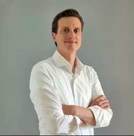 Persoon in wit overhemd met gekruiste armen tegen een grijze achtergrond.