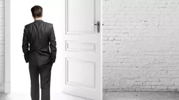 Person in a suit facing an open door in a bright room.