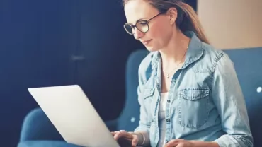 Person in denim shirt using a laptop on a couch.