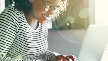 Person smiling while working on a laptop with sunlight streaming in.