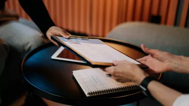 Twee personen wisselen een document uit boven een tafel.