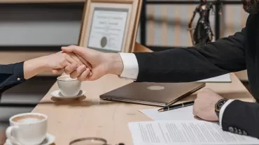 Twee handen schudden boven een tafel met papieren en een laptop.