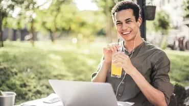 Person enjoying juice and listening to music on earphones, working on a laptop outdoors.