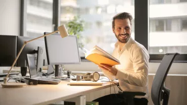 Persoon leest een boek op kantoor bij een bureau met computers.
