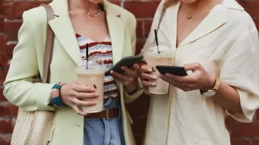 Two people holding iced coffees, looking at their phones against a brick wall.