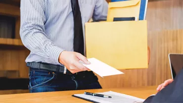 An employee, wearing a shirt and tie, extends a resignation letter to their boss while holding a box of documents.