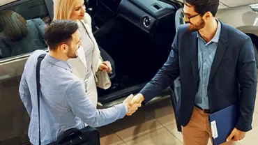 Three people standing by a car, shaking hands in a showroom.