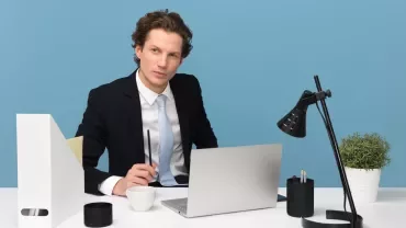Persoon achter bureau met laptop en notitieblok, blauwe achtergrond.