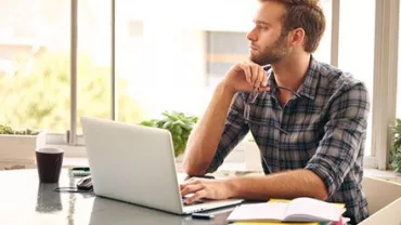 Person sitting at a table with a laptop, looking thoughtfully out the window.