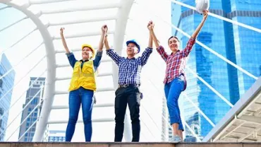 Three people in construction helmets cheering under modern architecture.