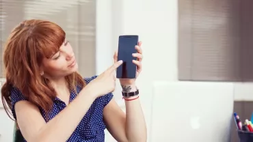 Persoon wijst naar mobiele telefoon bij een bureau met laptop.