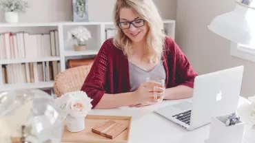 Persoon aan bureau met laptop en kopje thee, tussen boekenplanken.
