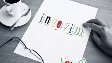 Hand holding paper with 'interim' in colorful letters, glasses, and coffee cup on table.