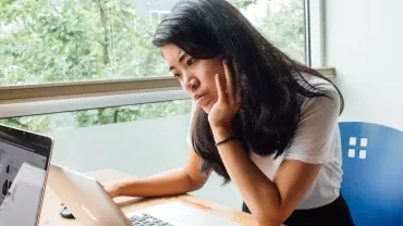 Person sitting at a desk, looking at a laptop near a window.
