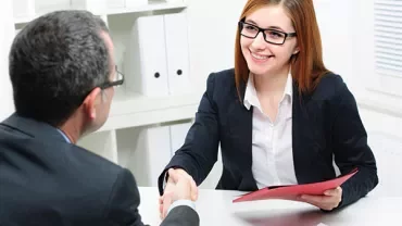 Two people shaking hands at a desk during a meeting.