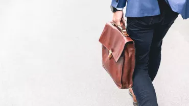 A person in a blue jacket walks with a brown briefcase.