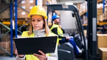 Person in warehouse checks clipboard; forklift in the background.