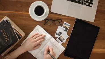 Person writing in notebook with laptop and coffee on table.