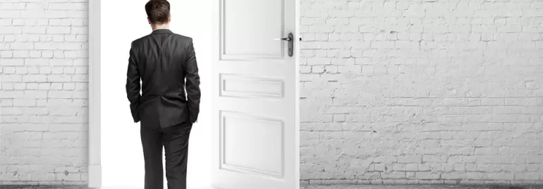 Person in a suit facing an open door in a bright room.