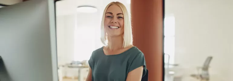 Vrouw die lacht achter een computerscherm in een kantooromgeving.