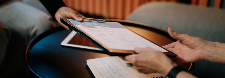 Twee personen wisselen een document uit boven een tafel.