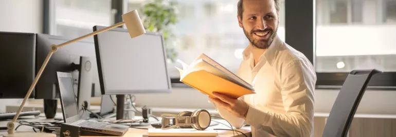 Persoon leest een boek op kantoor bij een bureau met computers.