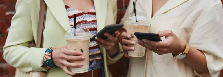 Two people holding iced coffees, looking at their phones against a brick wall.