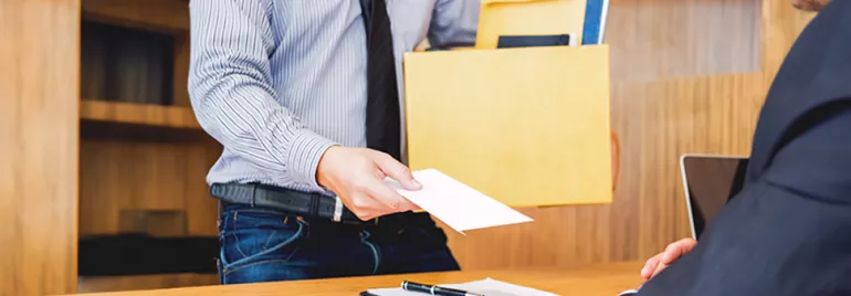 An employee, wearing a shirt and tie, extends a resignation letter to their boss while holding a box of documents.