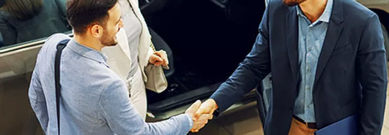 Three people standing by a car, shaking hands in a showroom.