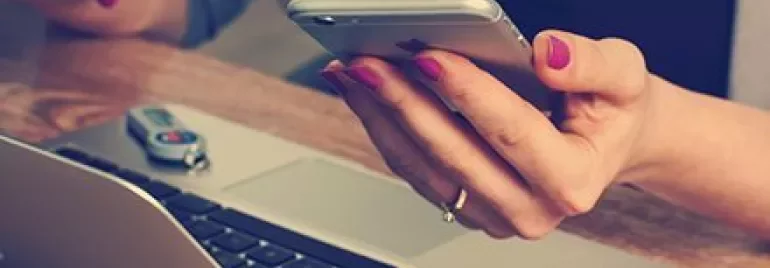 Handen met een telefoon en laptop op een bureau.