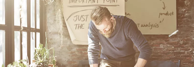 Man werkt aan bureau met papieren en kamerplanten, brainstormende aantekeningen op muur.
