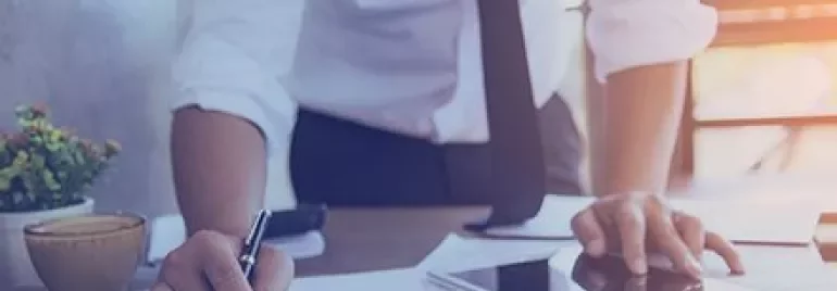 Person writing on documents beside a tablet and coffee.