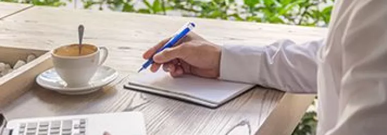 Person writing near a laptop with a cup of coffee on a wooden table.