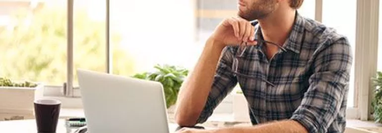 Person sitting at a table with a laptop, looking thoughtfully out the window.