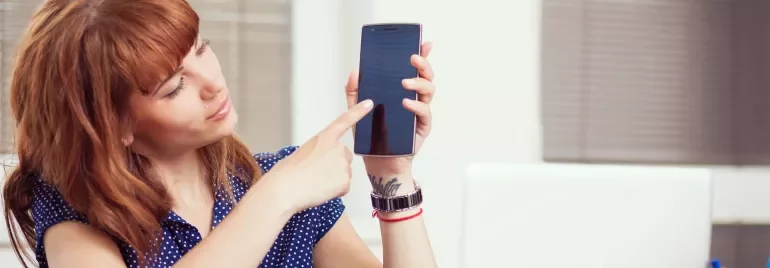 A person points at a phone while sitting at a desk with a laptop.