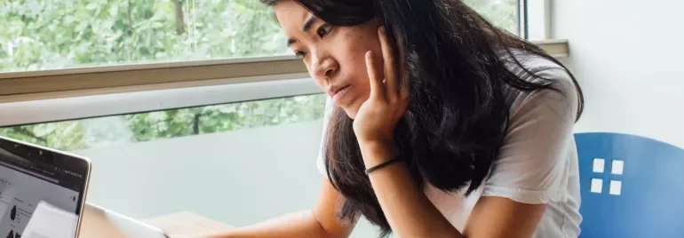 Persoon zittend aan een bureau, kijkend naar een laptop bij een raam.
