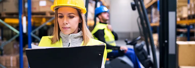 Person in warehouse checks clipboard; forklift in the background.