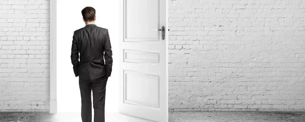 Person in a suit facing an open door in a bright room.