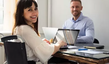 Twee mensen glimlachen en gebruiken laptops aan tafel in een kantoor.