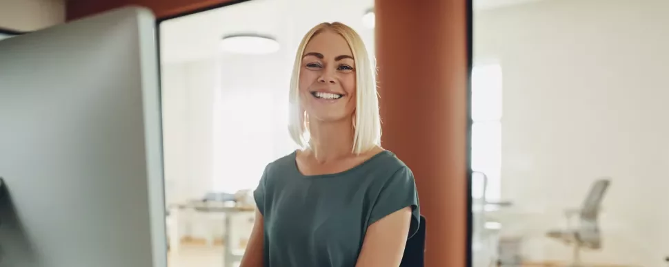 Vrouw die lacht achter een computerscherm in een kantooromgeving.