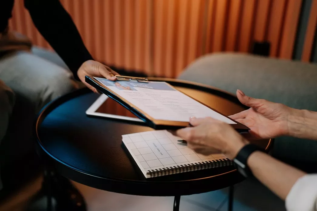 Twee personen wisselen een document uit boven een tafel.
