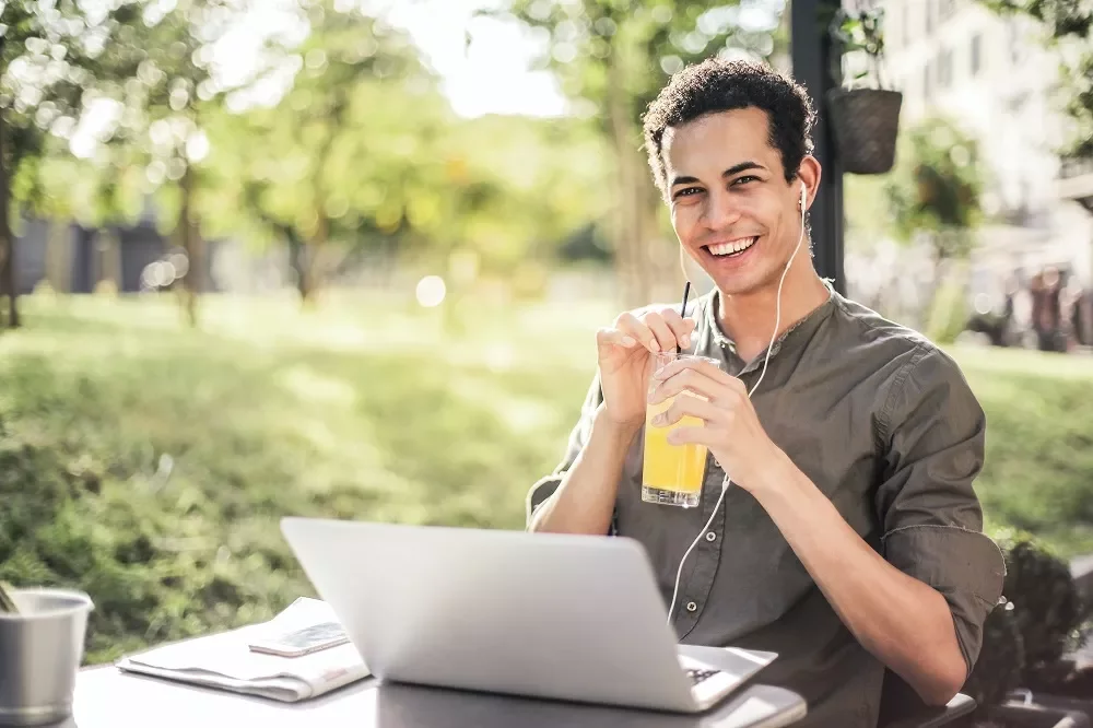 Person enjoying juice and listening to music on earphones, working on a laptop outdoors.