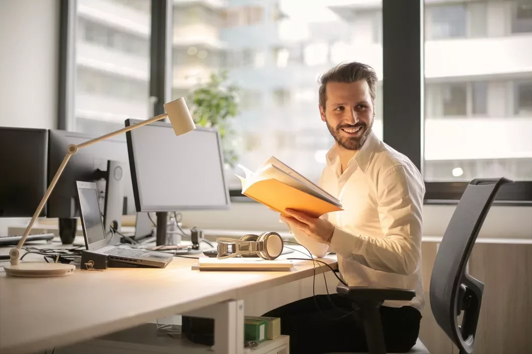 Persoon leest een boek op kantoor bij een bureau met computers.