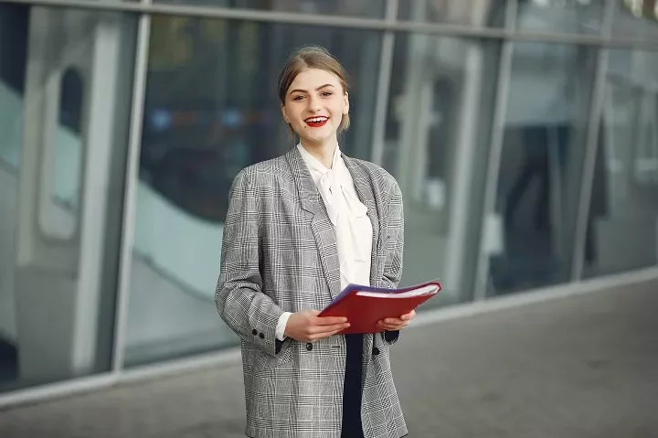Persoon met grijze blazer houdt rode map buiten een gebouw vast.