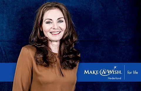Person smiling against Make-A-Wish Netherlands backdrop.