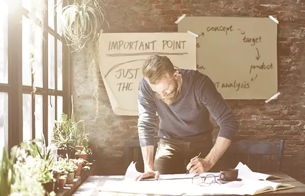 Man werkt aan bureau met papieren en kamerplanten, brainstormende aantekeningen op muur.