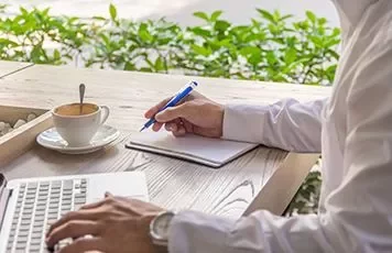 Persoon schrijft bij een laptop met een kopje koffie op een houten tafel.