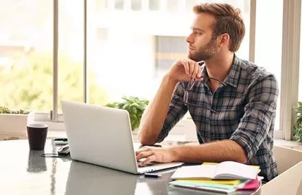 Person sitting at a table with a laptop, looking thoughtfully out the window.