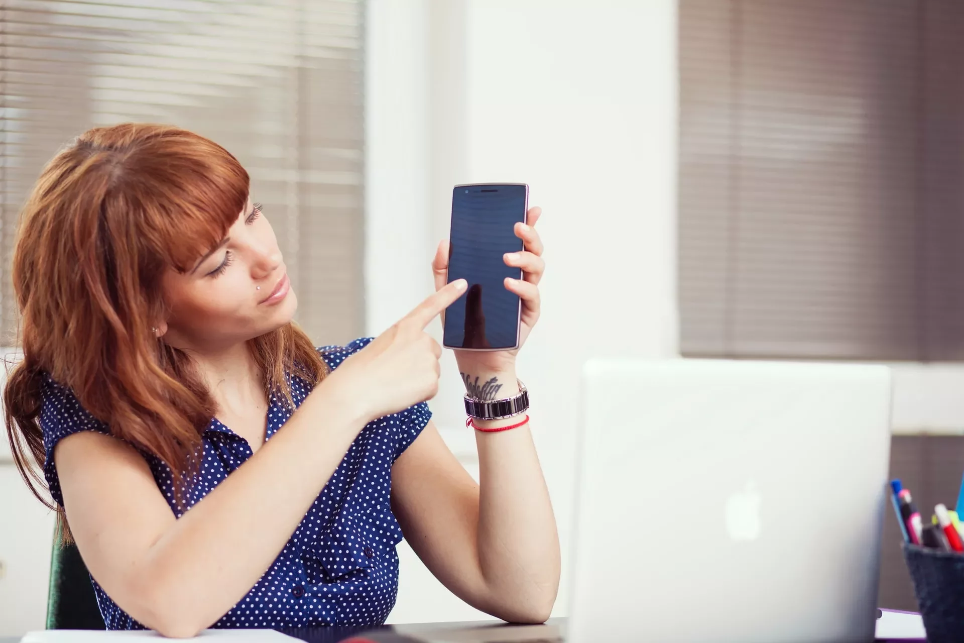 A person points at a phone while sitting at a desk with a laptop.